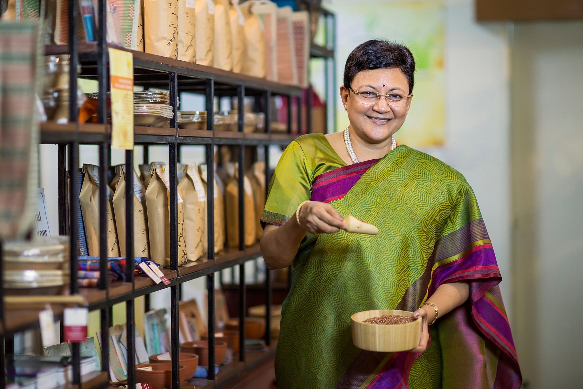 An Indian small-business owner in a green silk sari stands in her own retail store, holding a wooden bowl of grains, with shelves of packaged products behind her.